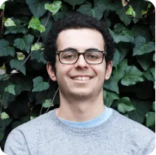 Young man with glasses and curly hair smiling in front of green leafy background.